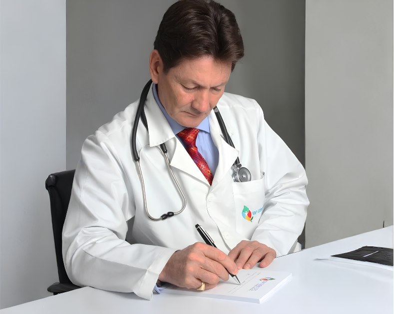 Dr. Van Uden firmando documentos en su consultorio, vestido con bata blanca y fonendoscopio